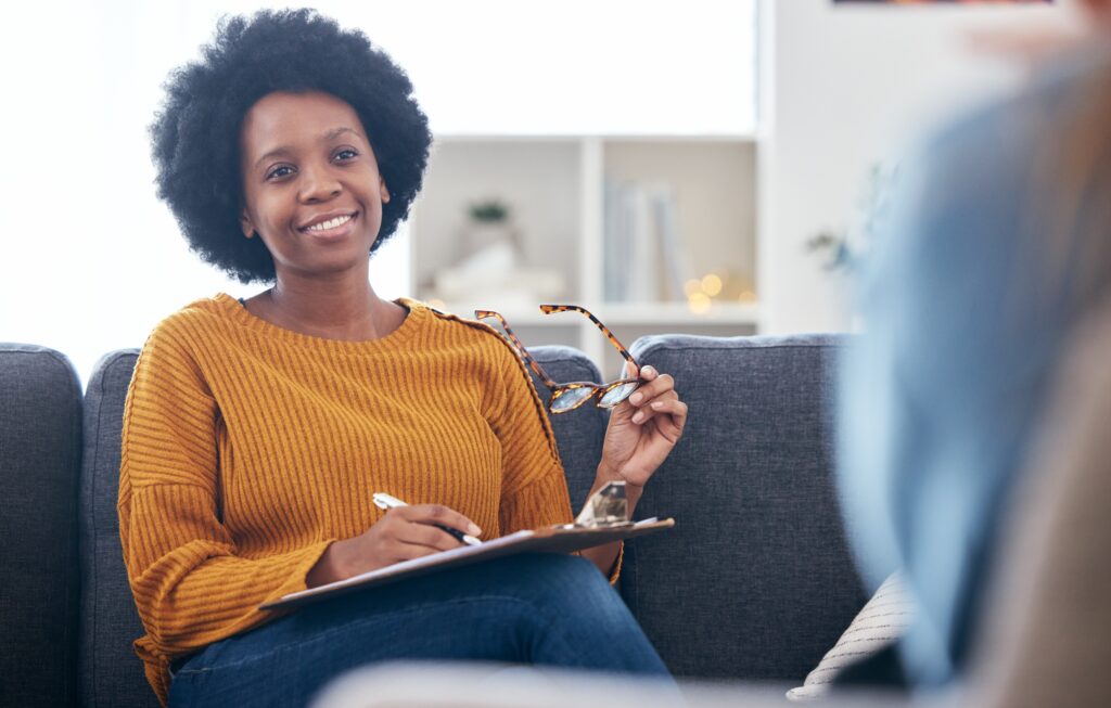 Psychologist on couch, writing notes with patient, advice and help in psychology, listening and mental health care. Conversation, support and black woman on sofa with client, therapist in counseling.