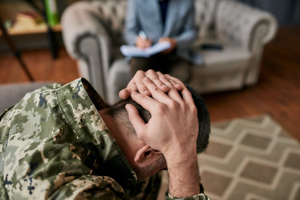 A man in military uniform stands with his head held in his hands, conveying a sense of distress or contemplation